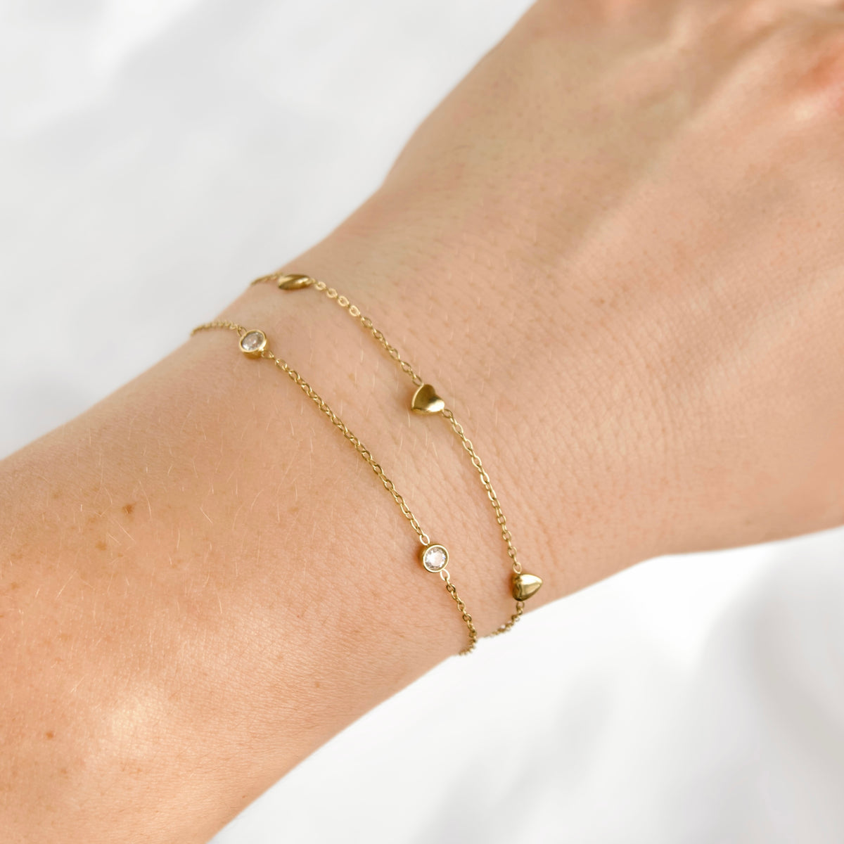 A close-up of a person's wrist wearing a layered delicate gold chain bracelet, one with small heart charms and one with a small round clear stone charm, against a soft white background.