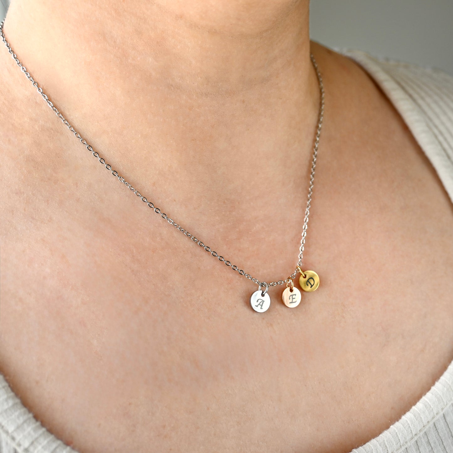 Three-toned necklace with three round initial pendants, one silver, one rose gold and one gold on a silver cable chain on a person wearing a light-colored top.