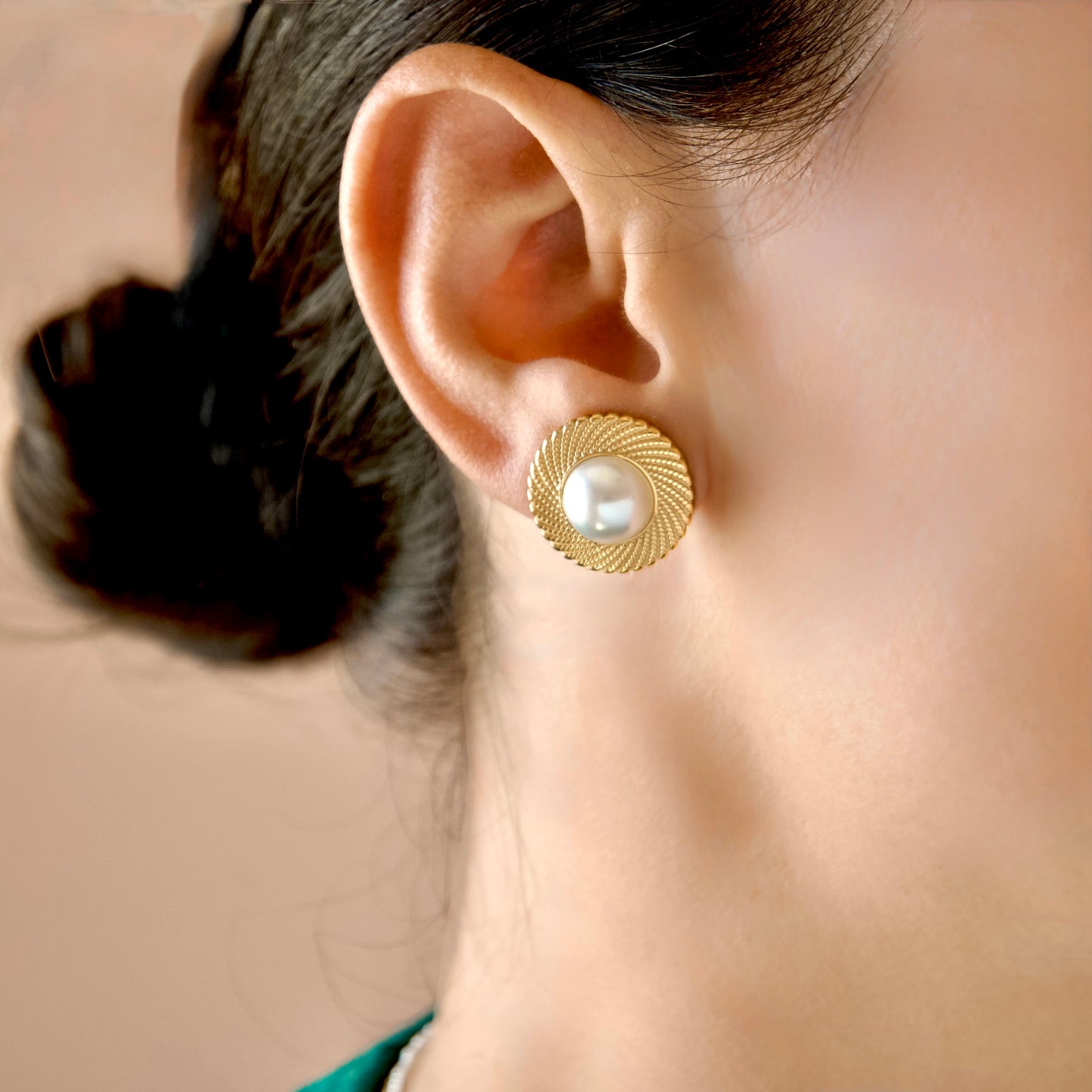 A close-up side profile of a woman wearing a gold textured circular stud earring with a white pearl center.