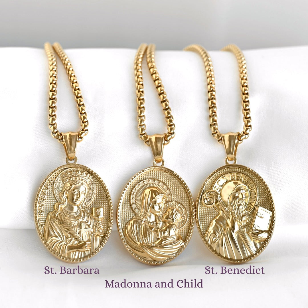 Three gold religious medallion necklaces displayed side by side on a white fabric surface. The necklaces feature oval pendants with embossed saint and religious imagery, labeled 'St. Barbara, Madonna and Child, and St. Benedict.