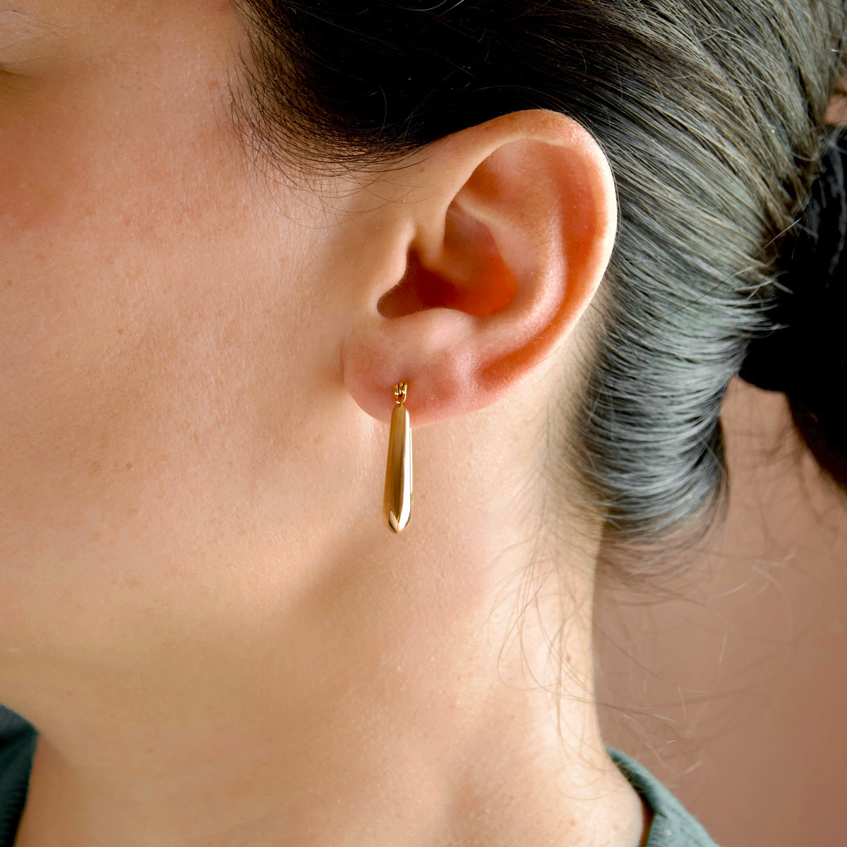 A close-up of a person's ear wearing a small, polished gold teardrop hoop earring.