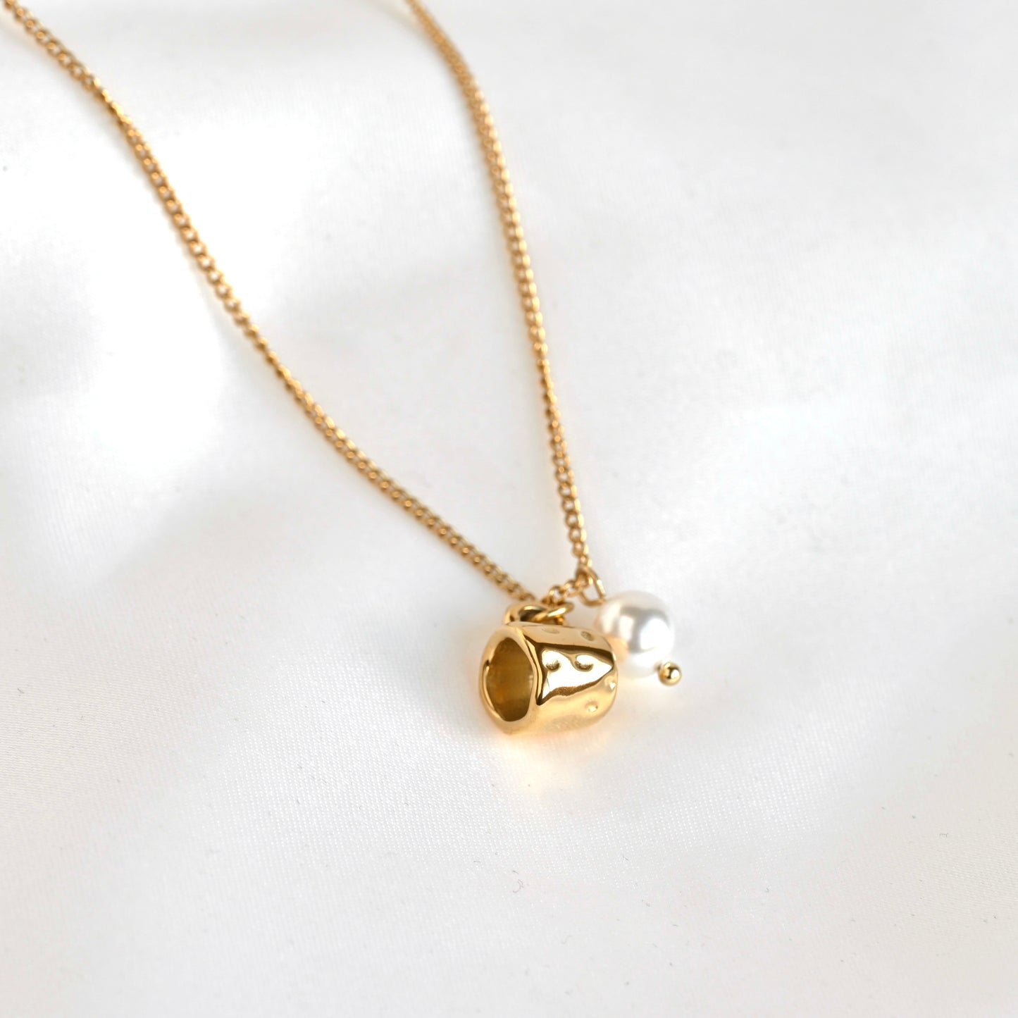 A gold chain necklace featuring a small gold pendant shaped like a cup, with a white pearl bead hanging beside it, displayed on a soft white fabric background.