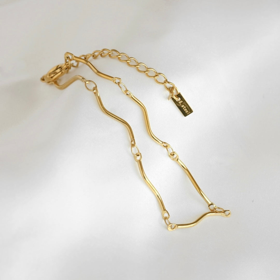 A gold-toned S-shaped link chain bracelet with an extender link and a small rectangular brand tag, photographed on a soft white fabric background.