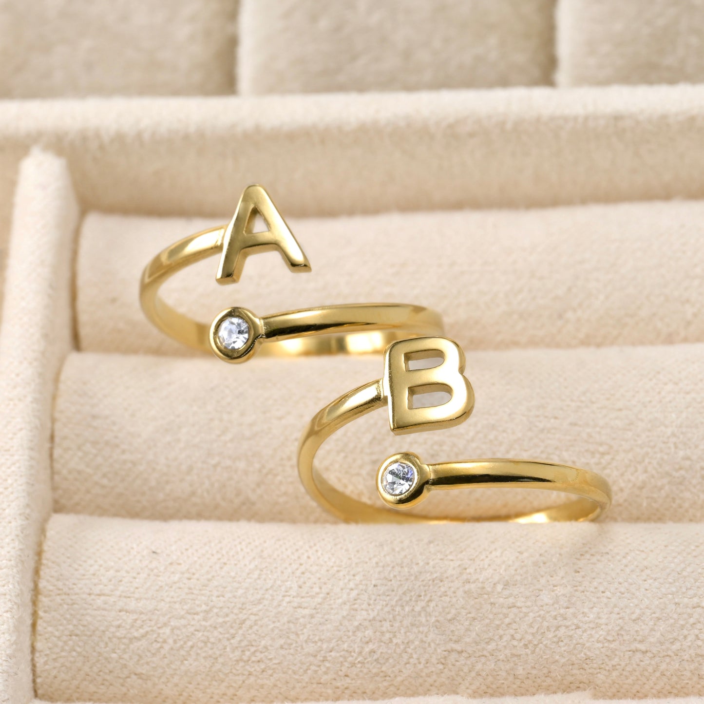Two dainty gold open rings with 'A' and 'B' letters on a beige fabric background