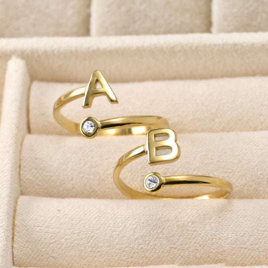Two dainty gold open rings with 'A' and 'B' letters on a beige fabric background