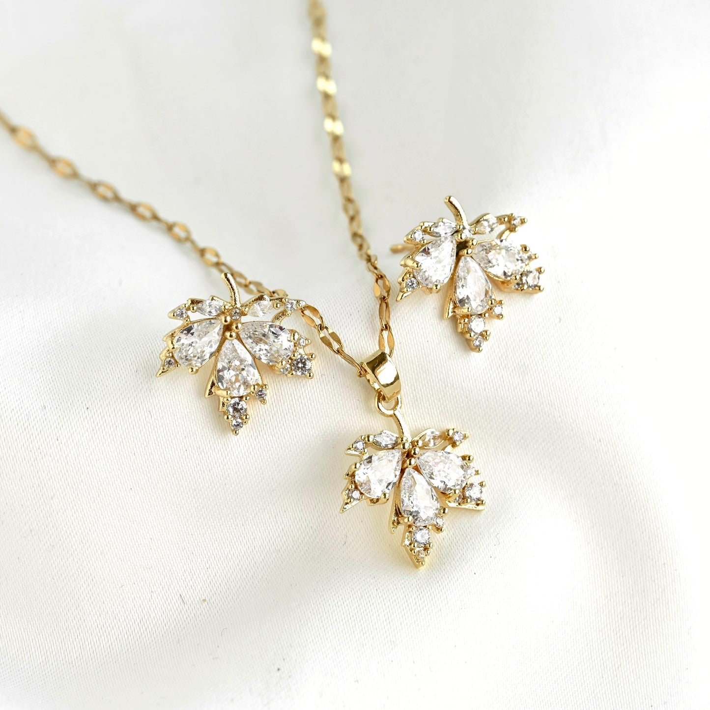 Gold leaf-shaped pendant necklace and matching earrings set with clear marquise-cut stones, displayed on a soft white fabric background.