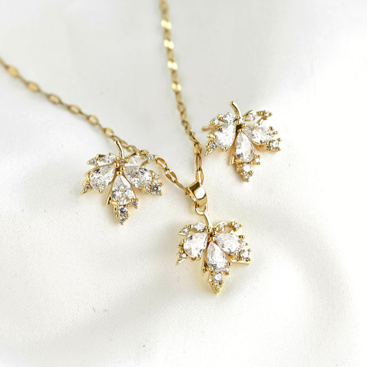 Gold leaf-shaped pendant necklace and matching earrings set with clear marquise-cut stones, displayed on a soft white fabric background.