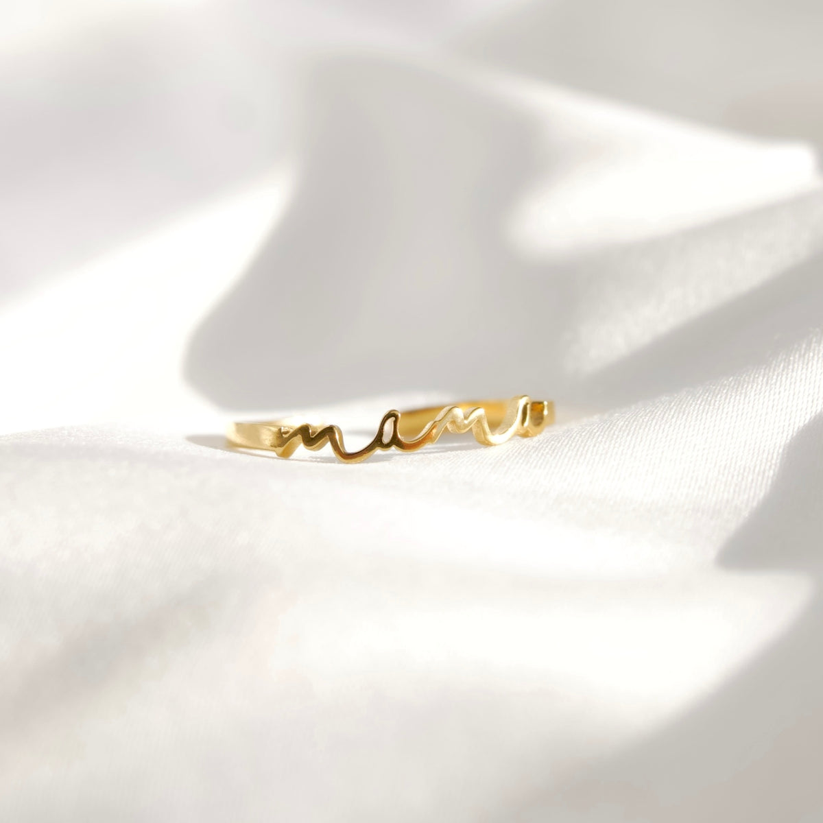 A delicate gold ring with a wavy script design spelling 'mama', resting on soft, draped white fabric with elegant shadows.