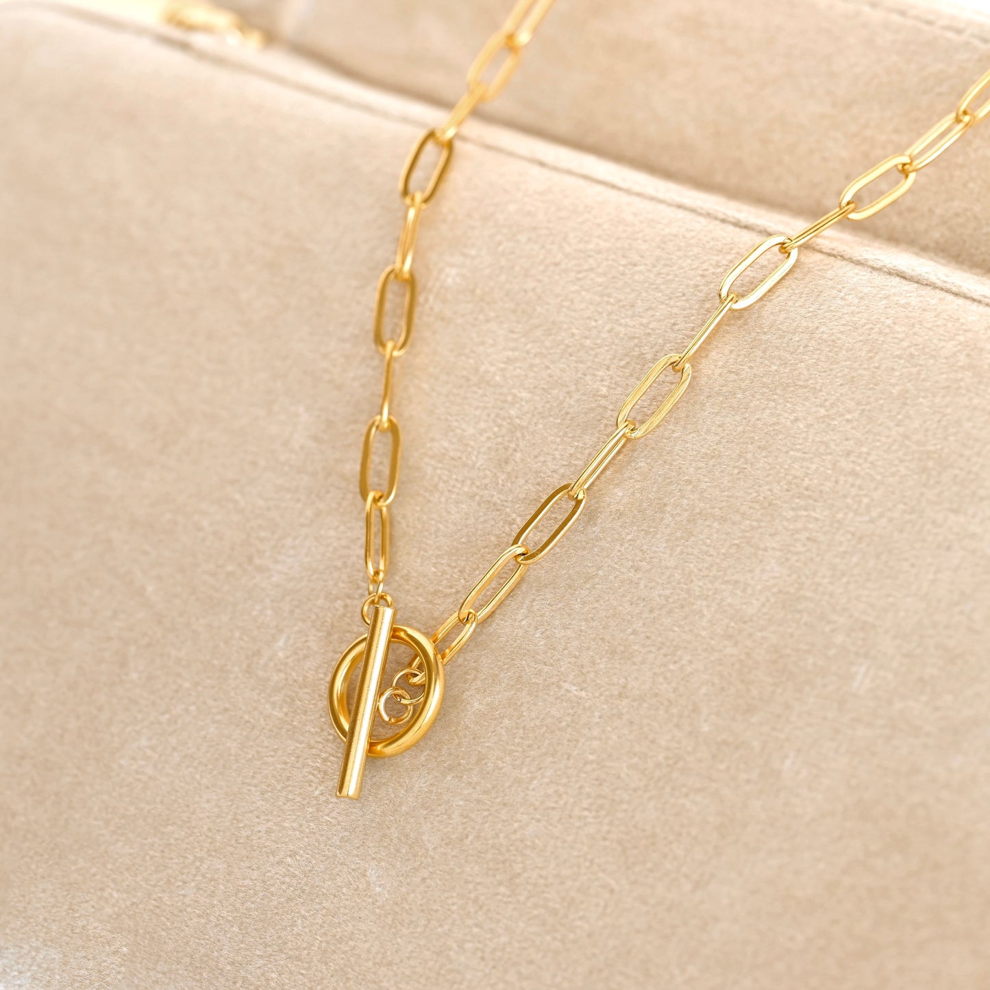 A gold paperclip chain necklace with a toggle clasp resting on a beige velvet jewelry display.