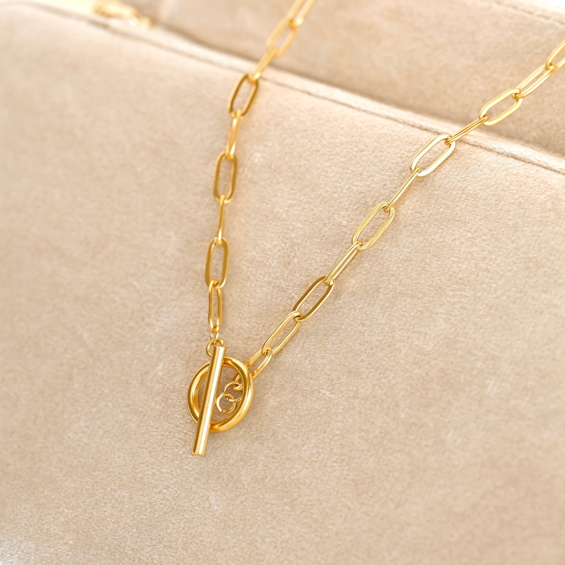 A gold paperclip chain necklace with a toggle clasp resting on a beige velvet jewelry display.