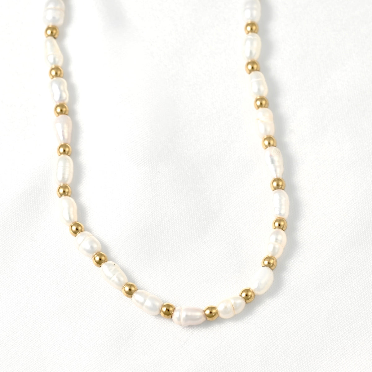 A delicate necklace featuring irregular white baroque pearls and small polished gold beads, arranged in a curved shape on a white fabric background.