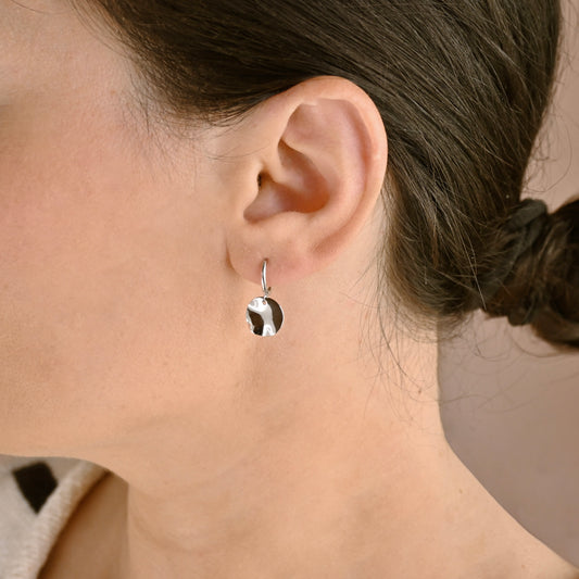 A close-up of a woman's ear wearing a small silver hoop earring with a dangling polished metal pendant shaped like a round leaf.