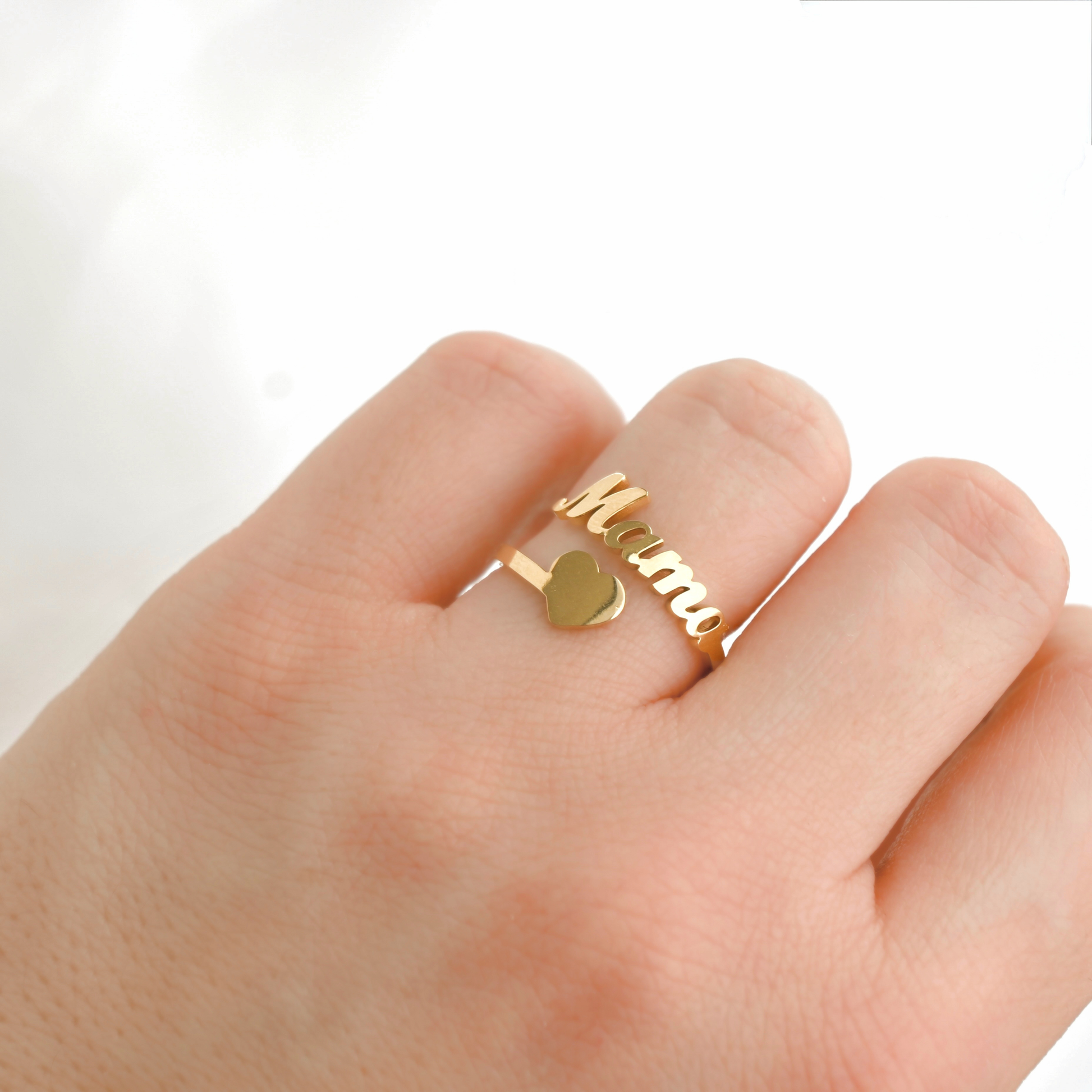 A gold-plated name ring with the word 'Mama' in cursive script and a small heart charm, shown worn on a person's finger against a bright white background.
