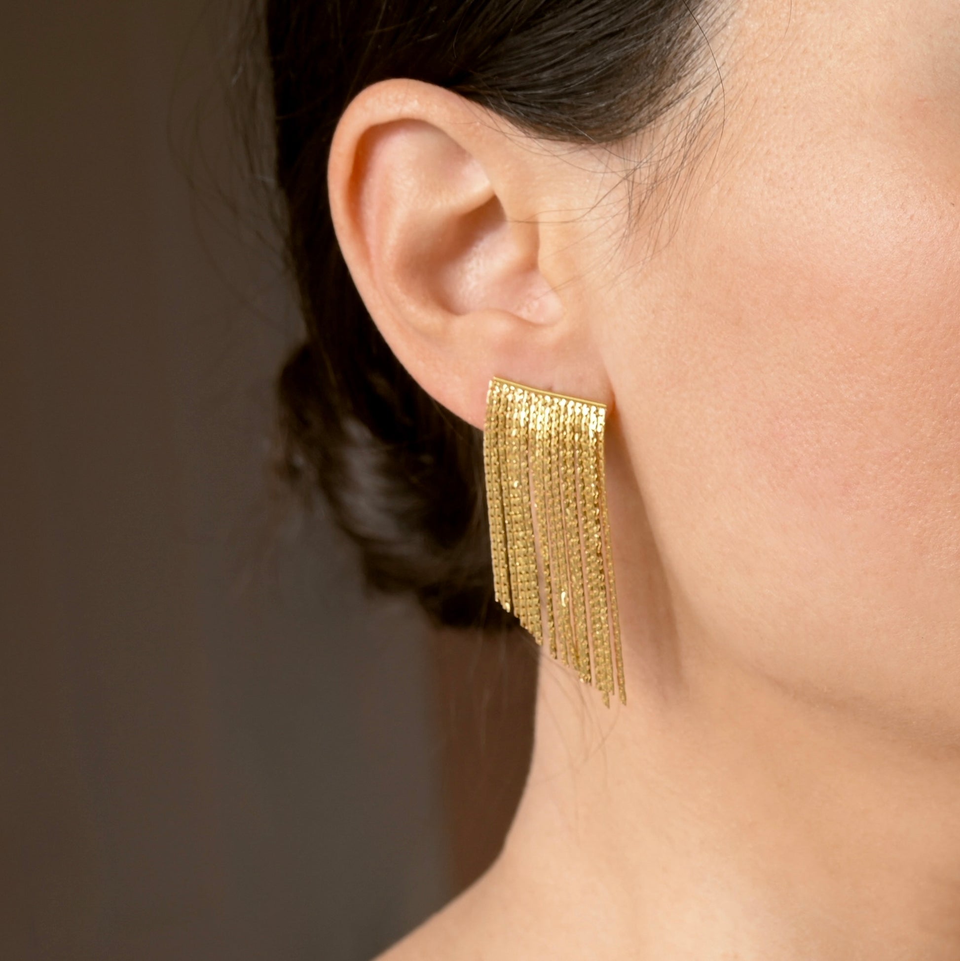 A close-up of a model's ear wearing a gold dangling earring with multiple cascading fringe strands.