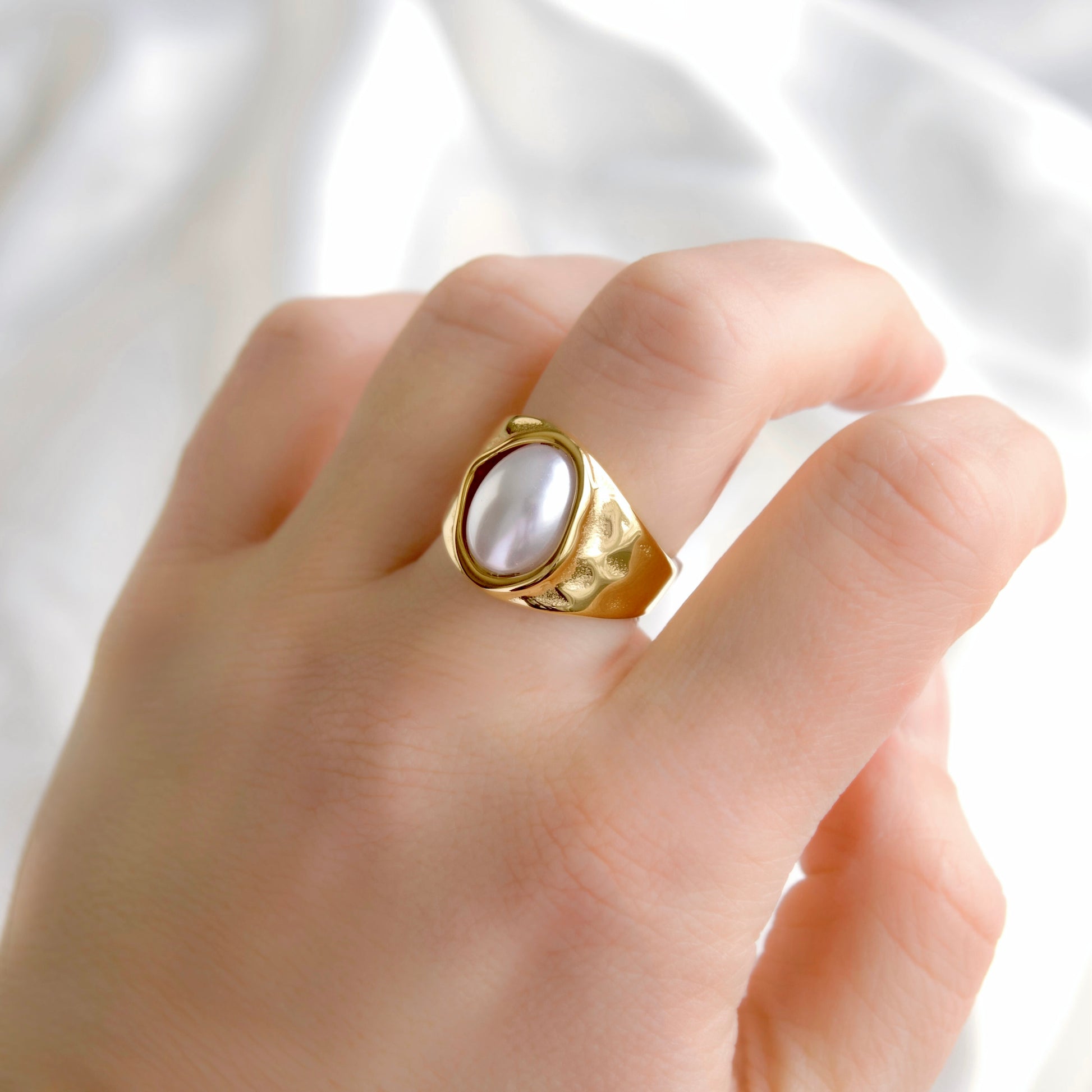 A gold statement ring with an oval white pearl-like center, worn on a person's finger against a soft white fabric background.