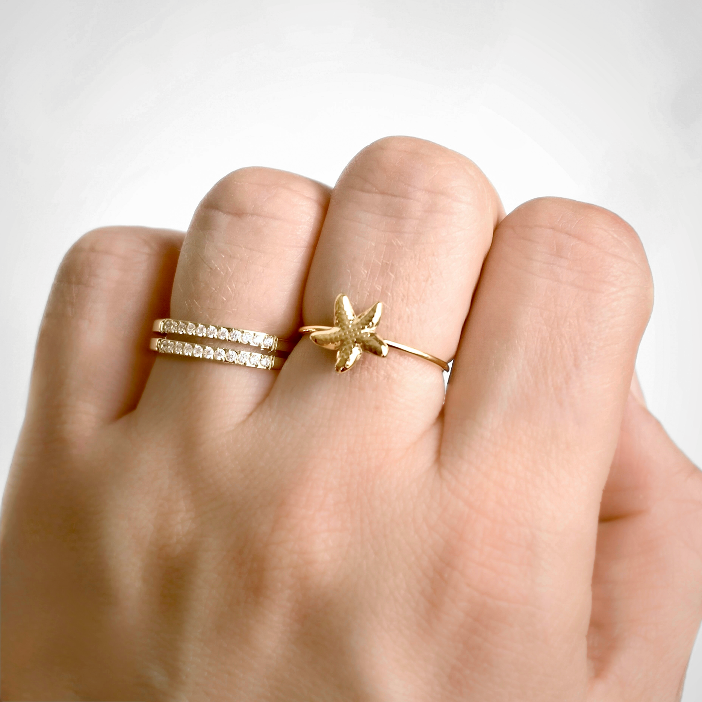 A close-up of a hand wearing two gold rings: a thin diamond-accented band on the ring finger and a textured starfish-shaped ring on the middle finger, against a clean white background.