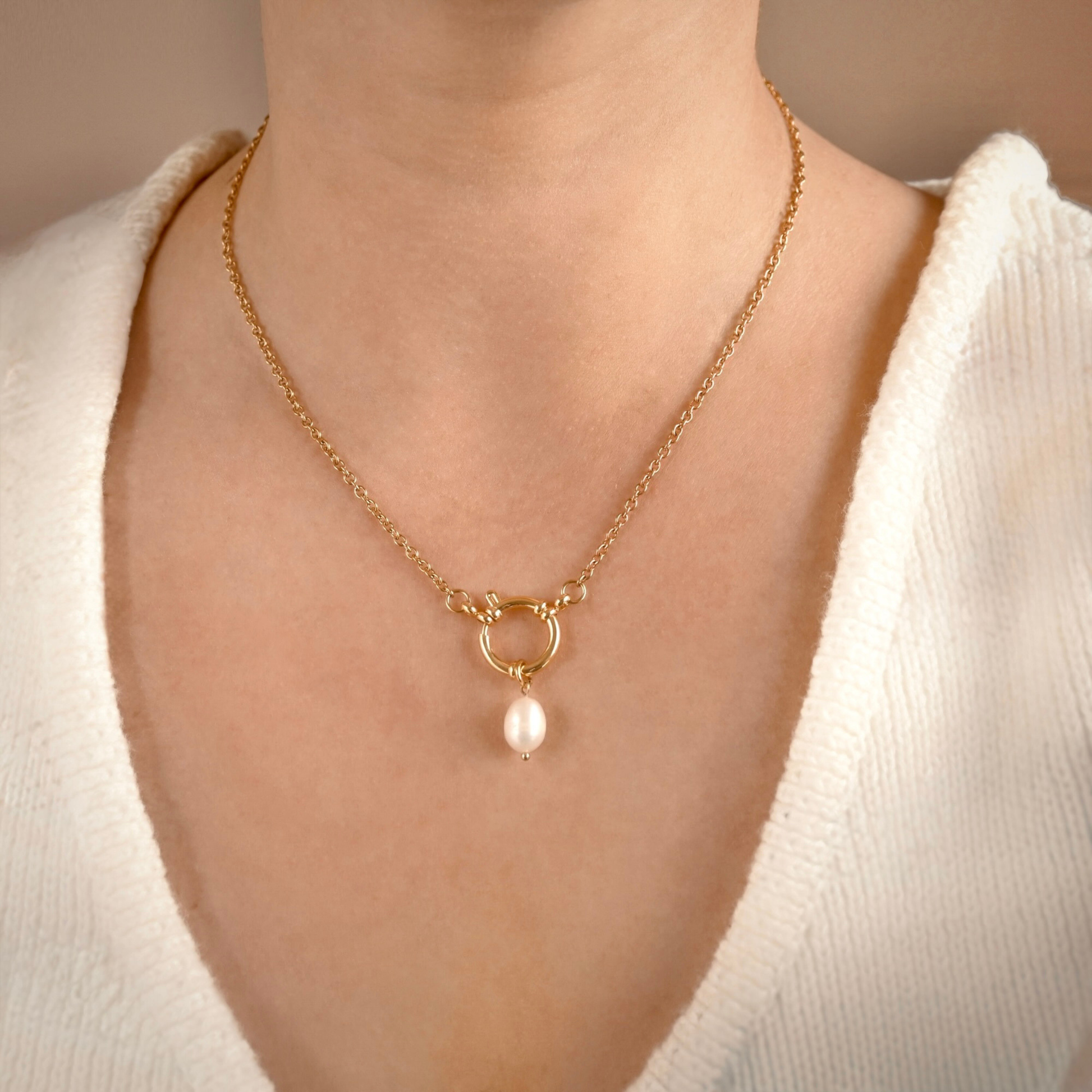 A close-up of a model wearing a delicate gold chain necklace with a front spring ring clasp and a single dangling white pearl pendant, styled with a white knit top.