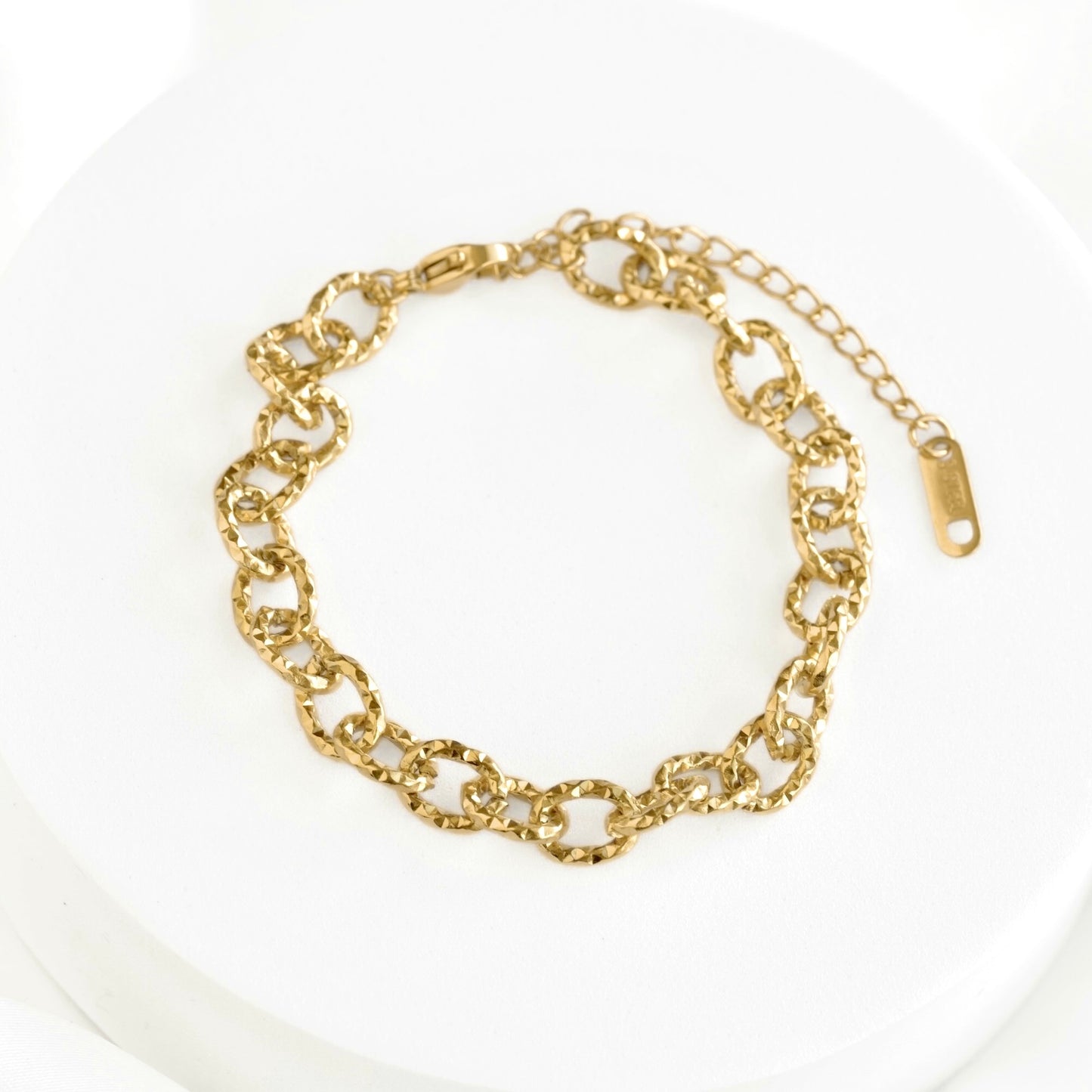 A gold-toned chain link bracelet with a lobster clasp and extender chain, displayed on a white circular surface.