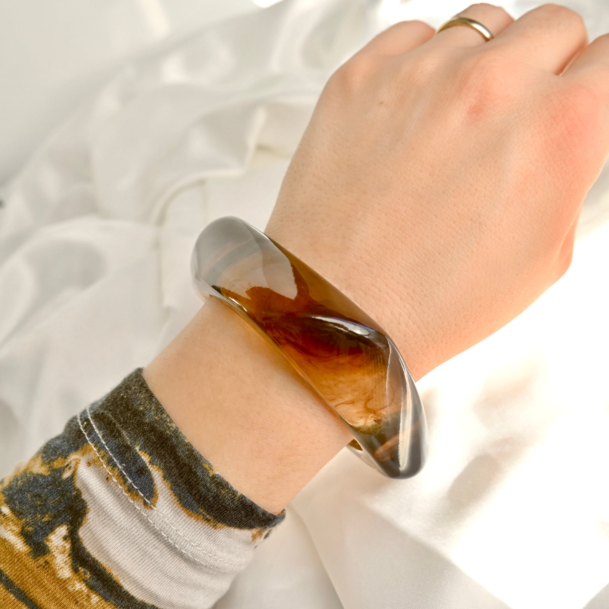 A close-up of a person's wrist wearing a chunky, polished brown and smoky gray bangle bracelet, with a patterned knit sleeve visible on the lower arm against a soft white fabric background.
