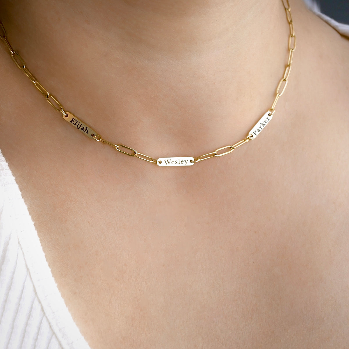 A close-up of a gold paperclip chain necklace worn on a person's neck, featuring three engraved name plates reading Elijah, Wesley, and Wesley.