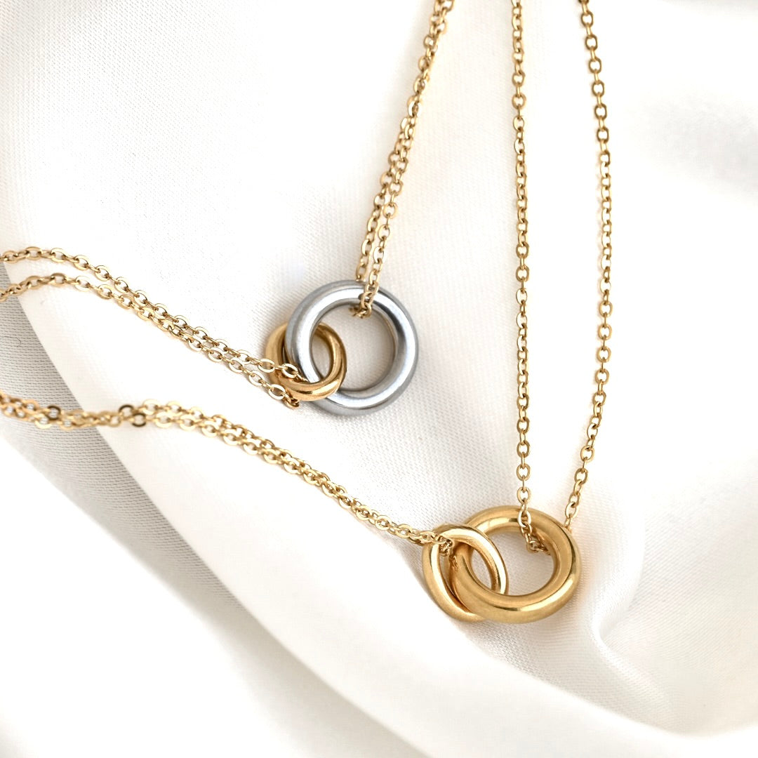 Two gold-toned interlocking ring necklaces with delicate chains, displayed on a soft white fabric background.