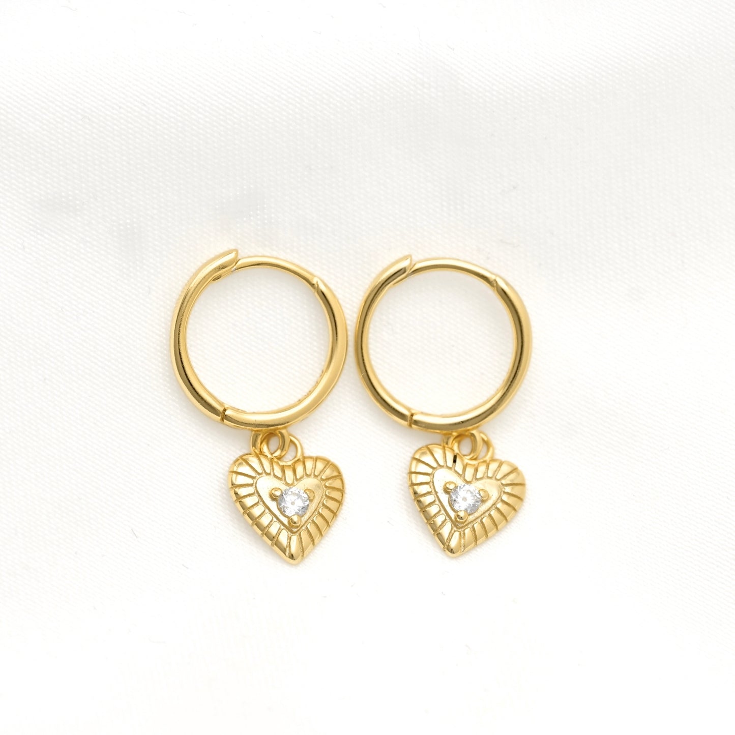 A pair of gold hoop earrings with small textured heart charms set with clear stones, displayed on a white textured fabric background.