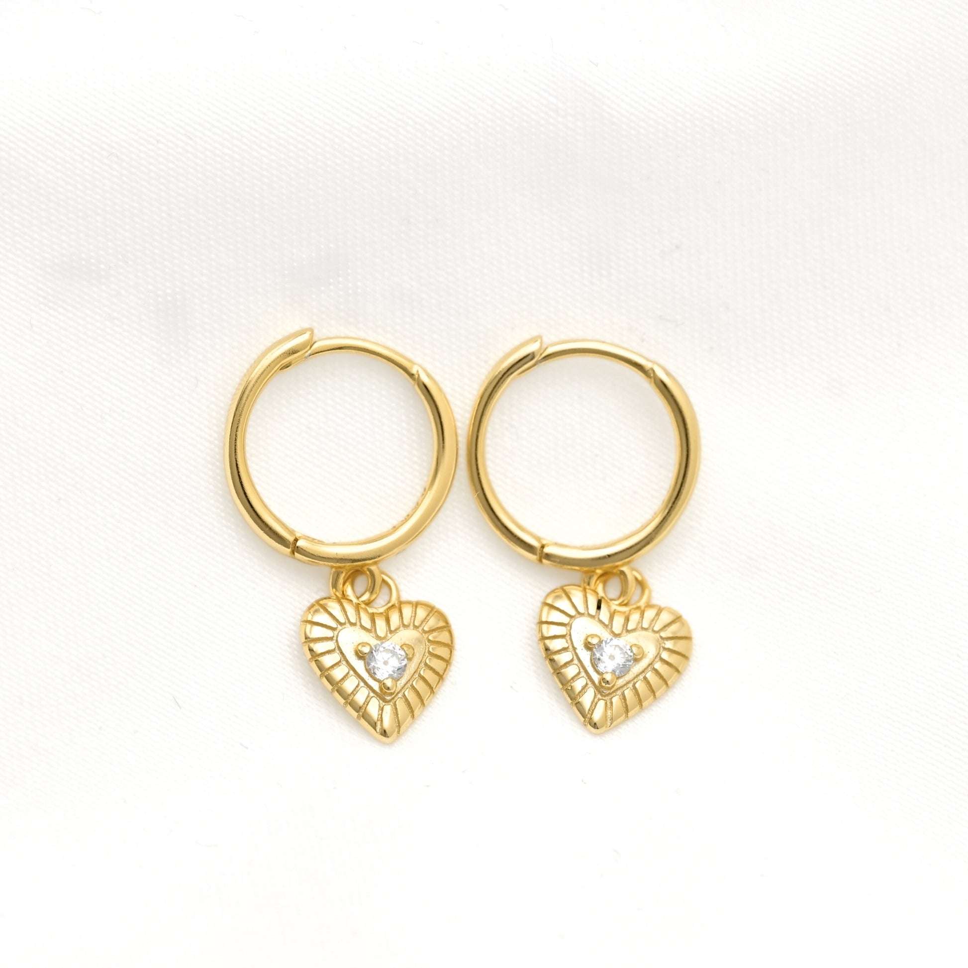 A pair of gold hoop earrings with small textured heart charms set with clear stones, displayed on a white textured fabric background.