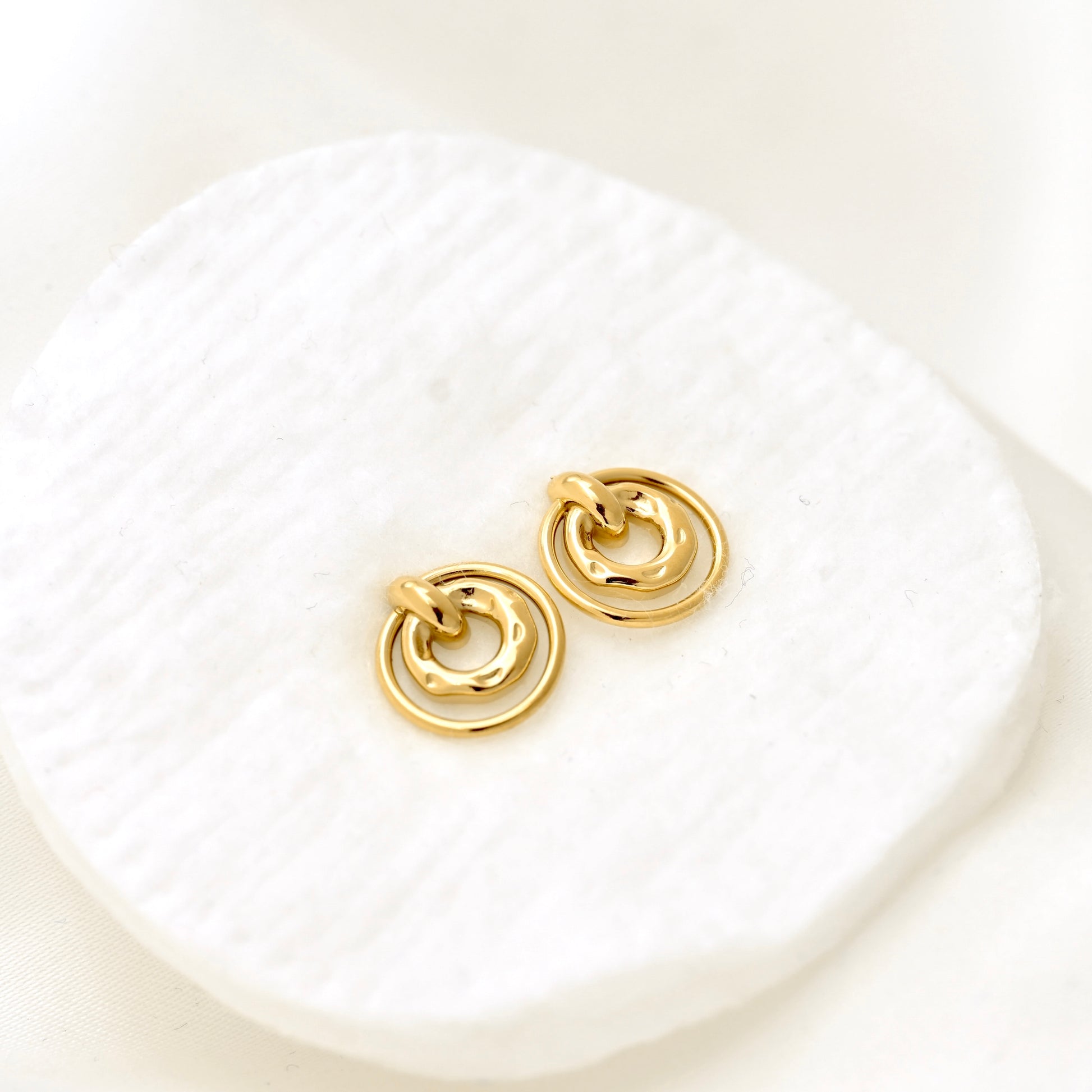 A pair of gold-toned double-ring hoop earrings resting on a textured white display pad against a soft white background.