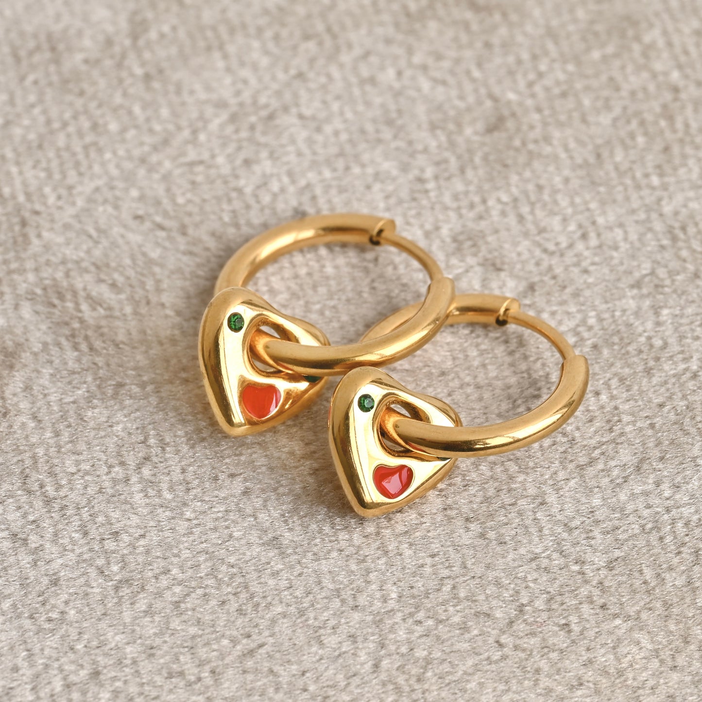 A pair of gold charm earrings with red and green stones on a beige velvet background