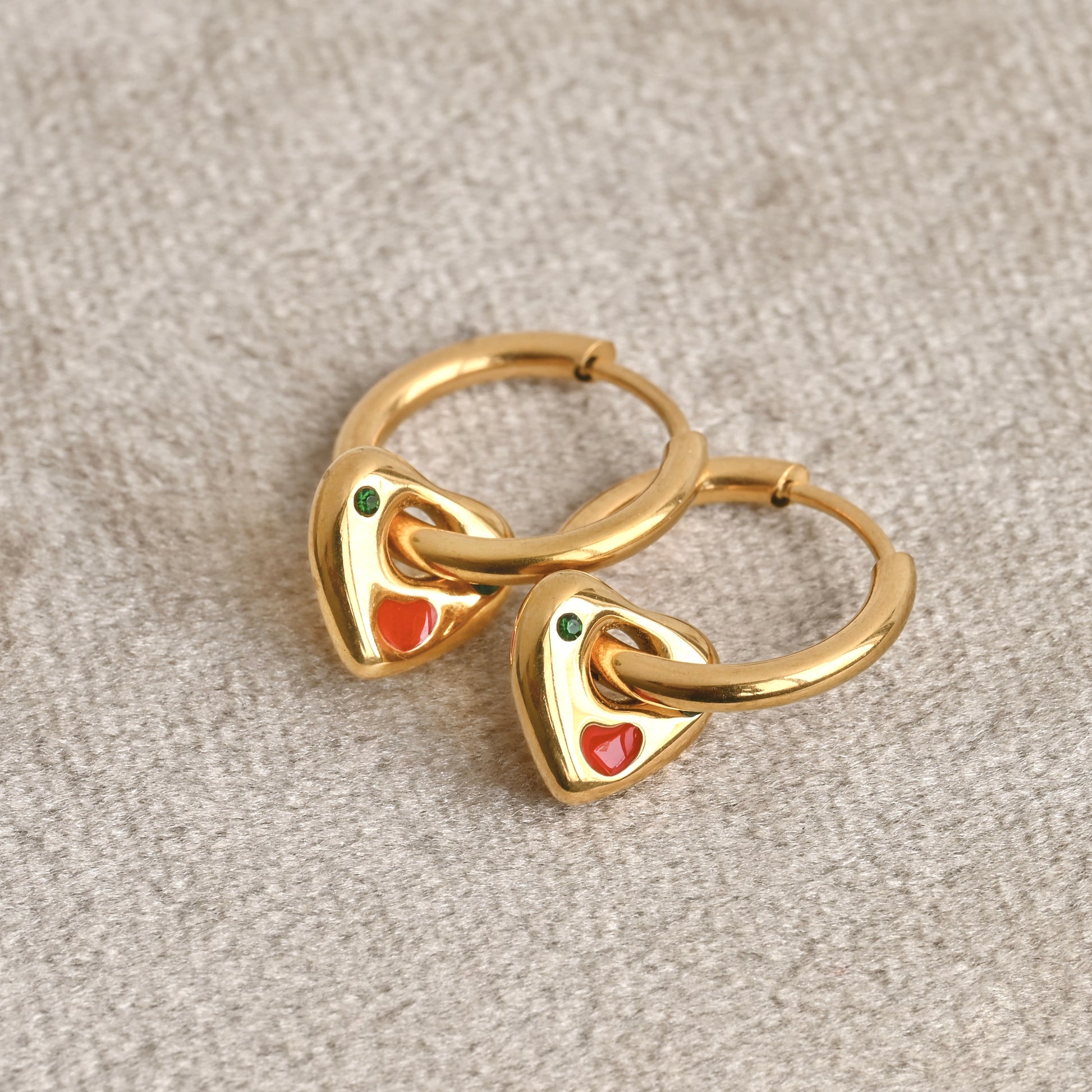 A pair of gold charm earrings with red and green stones on a beige velvet background