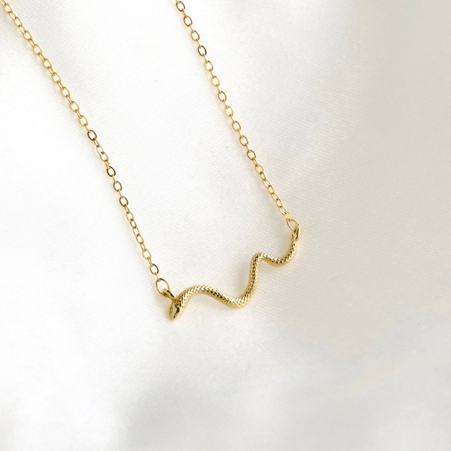A delicate gold chain necklace featuring a small, textured wavy pendant, displayed on a soft, white satin fabric background.