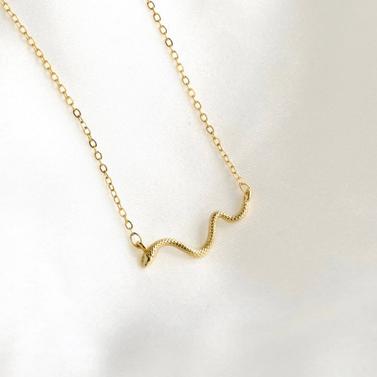 A delicate gold chain necklace featuring a small, textured wavy pendant, displayed on a soft, white satin fabric background.