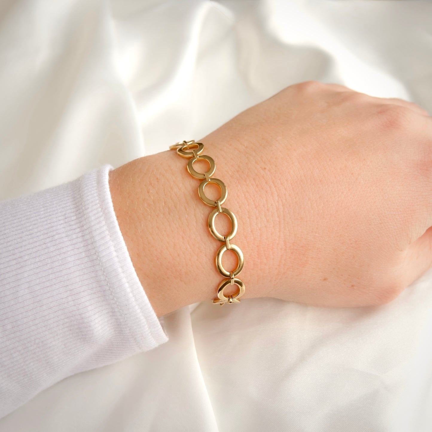A gold link bracelet with interlocking circular links worn on a person's wrist, shown against a soft white fabric background.