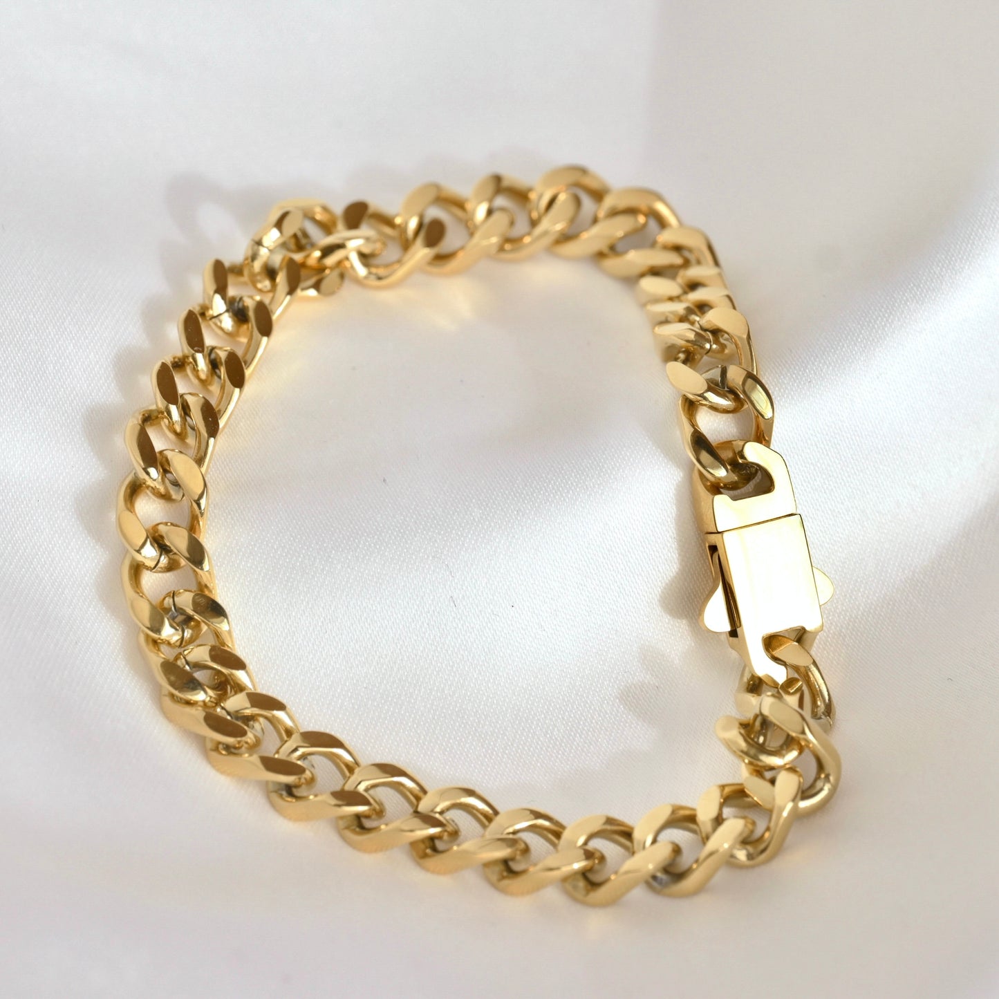 A gold-tone Cuban link chain bracelet with a polished box clasp, displayed on a white satin fabric background.