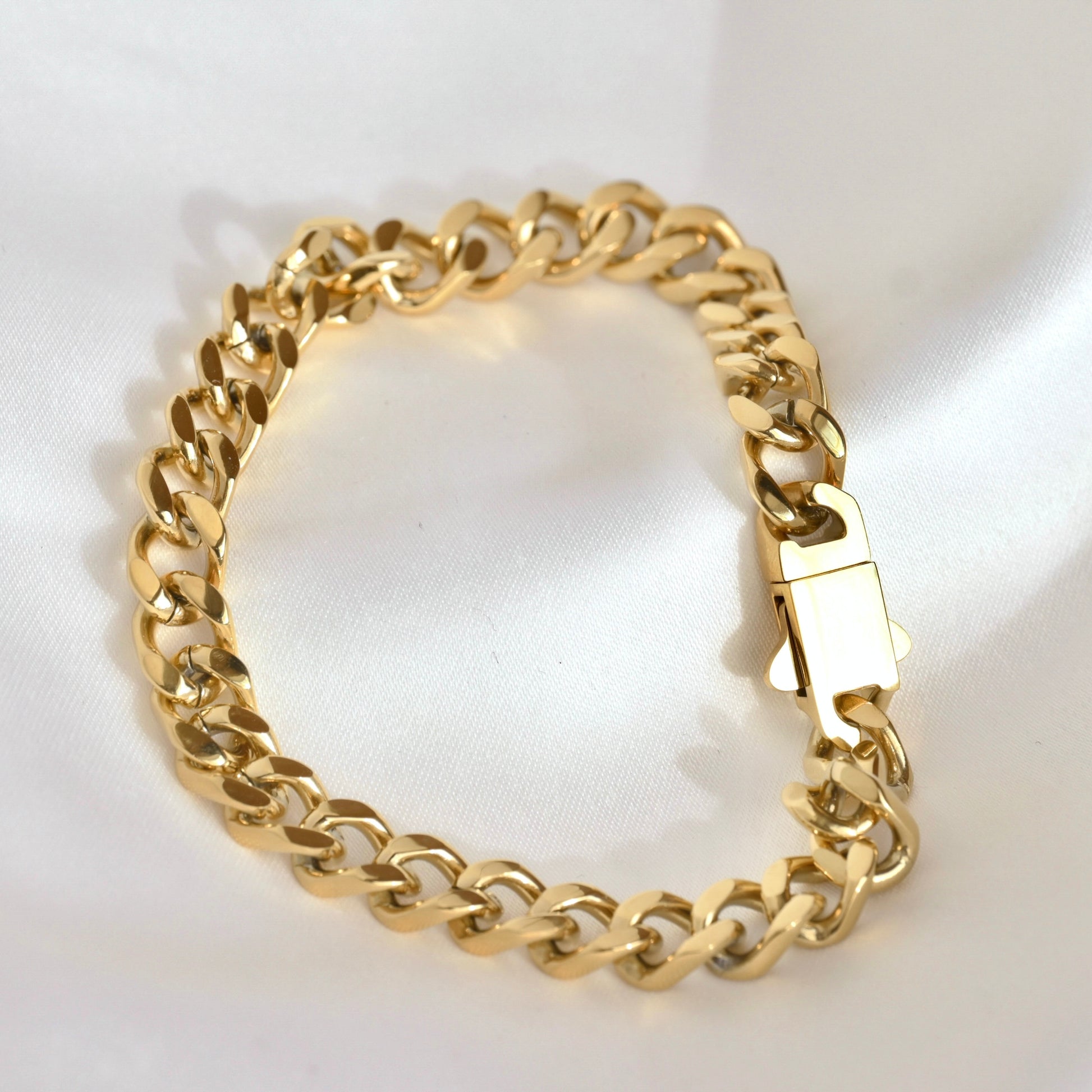 A gold-tone Cuban link chain bracelet with a polished box clasp, displayed on a white satin fabric background.