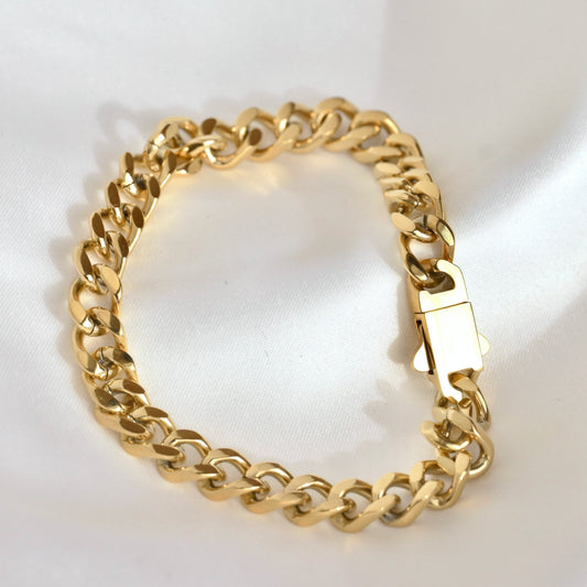 A gold-tone Cuban link chain bracelet with a polished box clasp, displayed on a white satin fabric background.