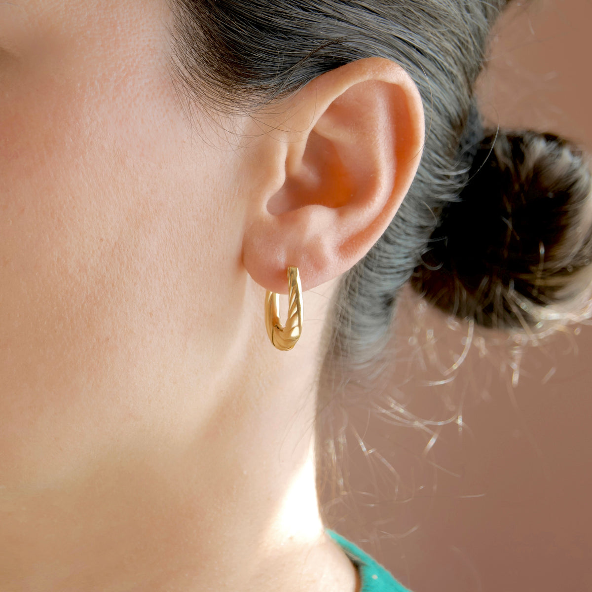 A close-up side profile of a woman wearing a small gold huggie hoop earring. Her hair is pulled back into a bun, and the background is a soft, warm neutral color.