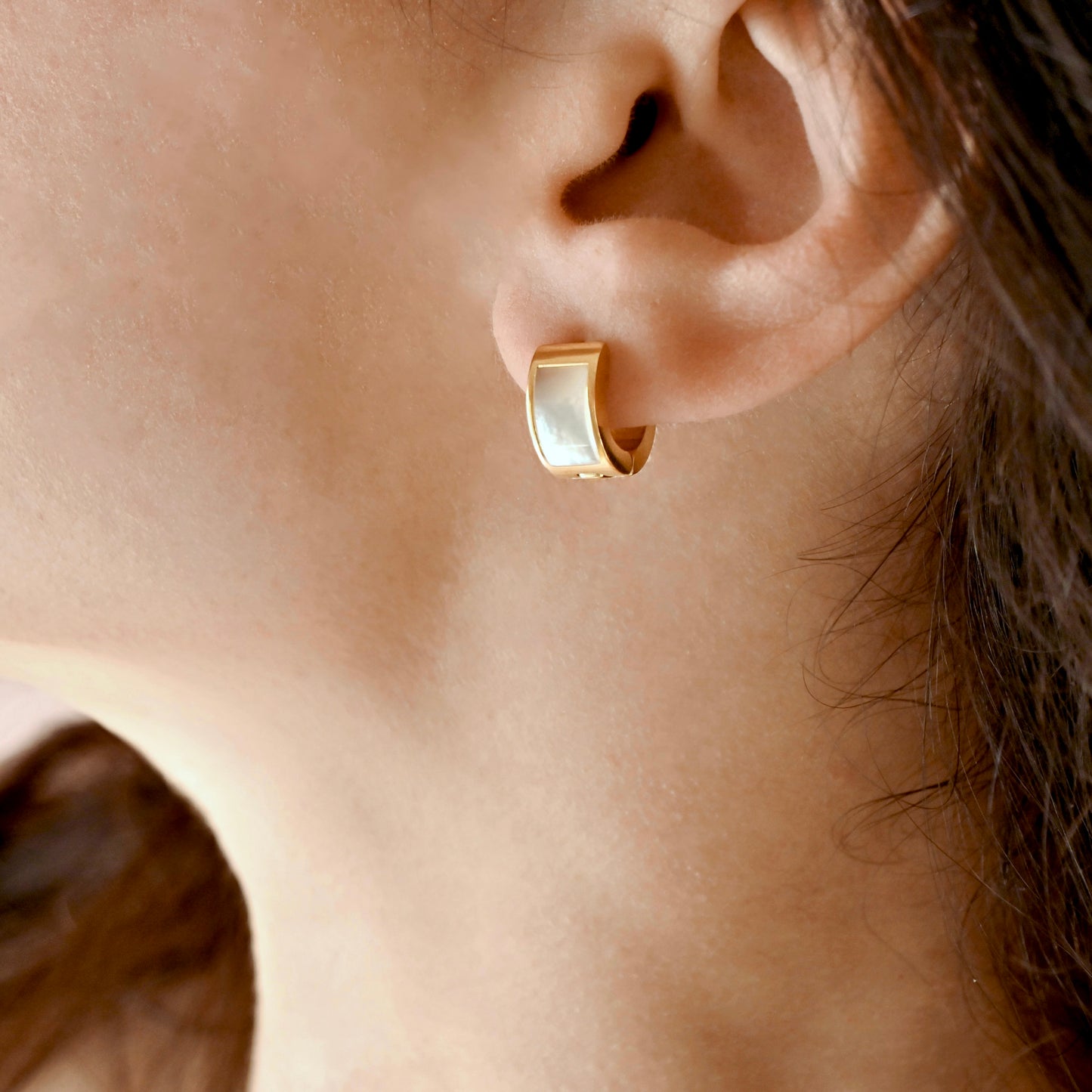 A close-up of a person's ear wearing a small gold huggie hoop earring with a smooth, polished white mother-of-pearl inlay.
