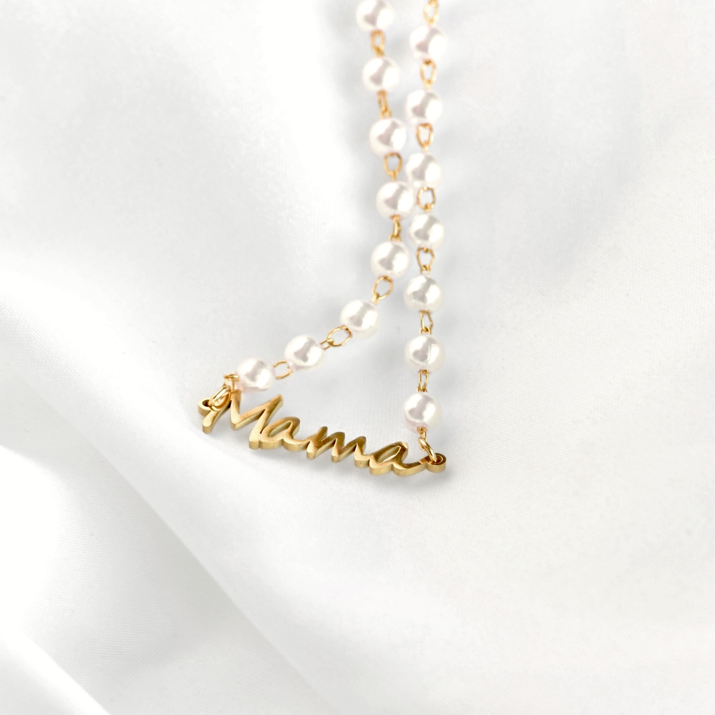 A delicate necklace featuring white faux-pearl beads on a gold chain, with a gold script pendant spelling 'Mama', displayed on a soft white fabric background.