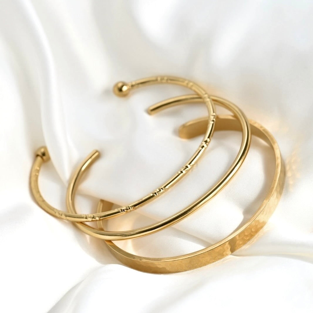 Three gold-toned open cuff bracelets with varying textures, displayed on a soft white satin fabric background.
