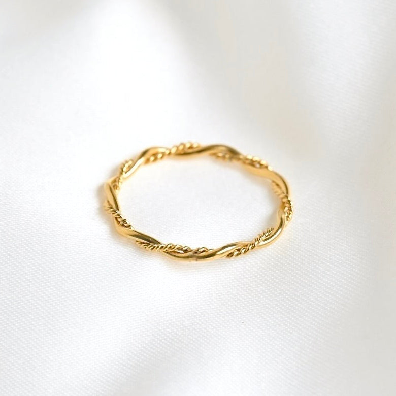 A delicate gold ring with a twisted, rope-like textured band, resting on a soft, white fabric background.