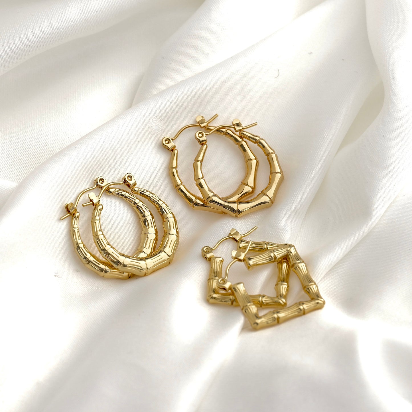 Three pairs of gold hoop earrings with a textured bamboo or segmented branch design, two round and one square designs, arranged on a draped white satin fabric background.