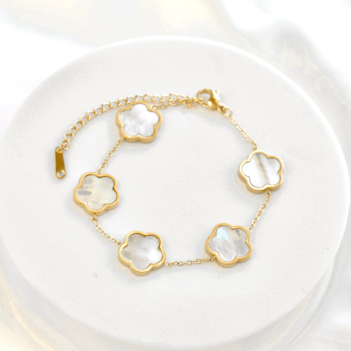 A gold chain bracelet with five flower-shaped mother-of-pearl links, displayed on a white circular stand.