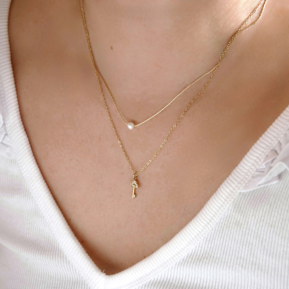 A close-up of a person's neck and chest wearing two delicate gold necklaces layered over a white ribbed top. The shorter chain has a small pearl bead, and the longer chain has a small gold key pendant.