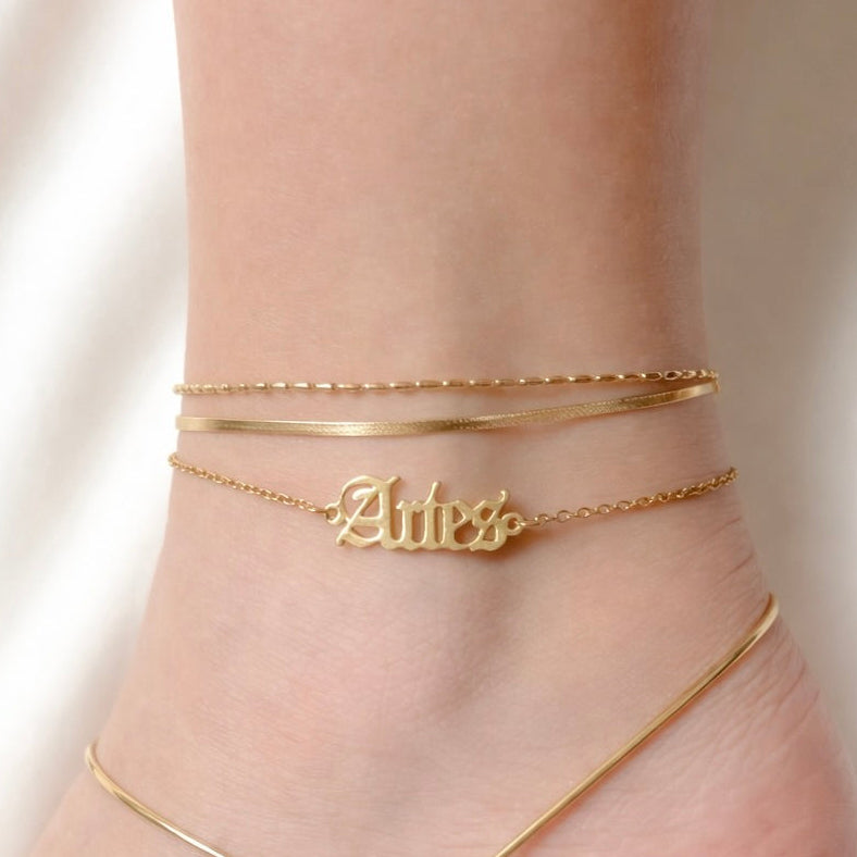 Gold anklets stacked on an ankle, one double layered and one with 'Aries' script with a plain background