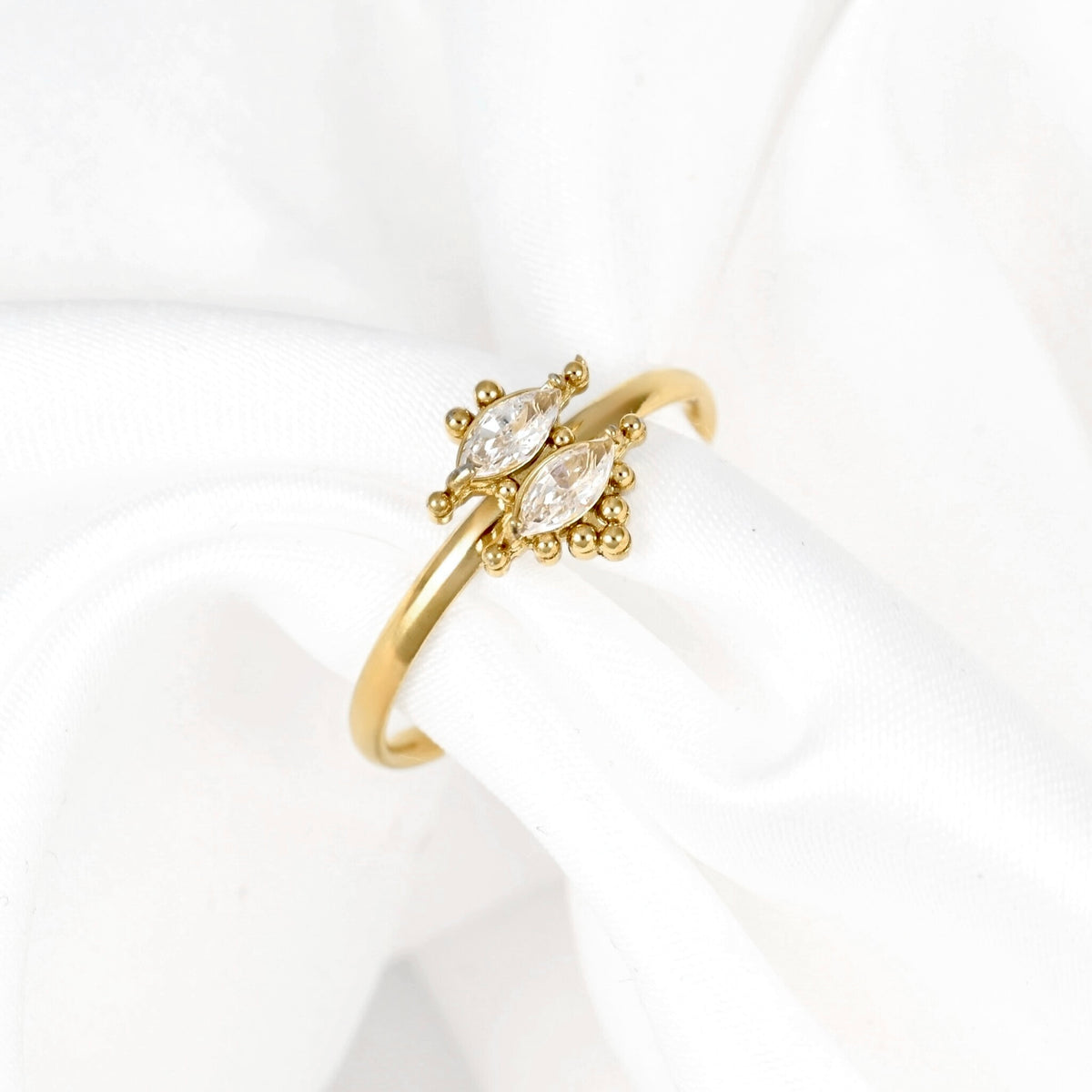 A gold ring with two marquise-cut clear gemstones and small gold accents, displayed on a white fabric ring stand.