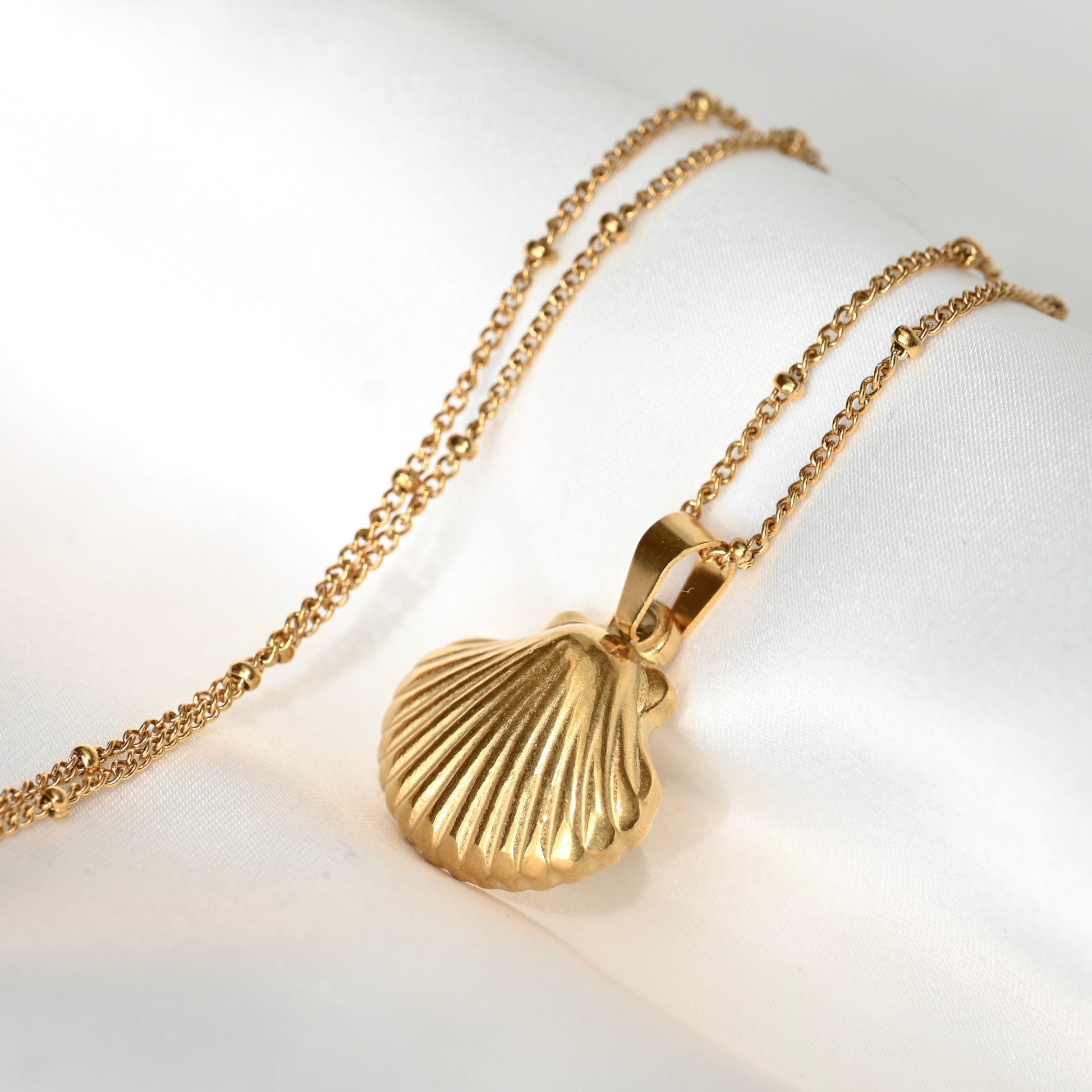 A gold shell pendant necklace  with a thin satellite chain displayed on a white satin fabric background.