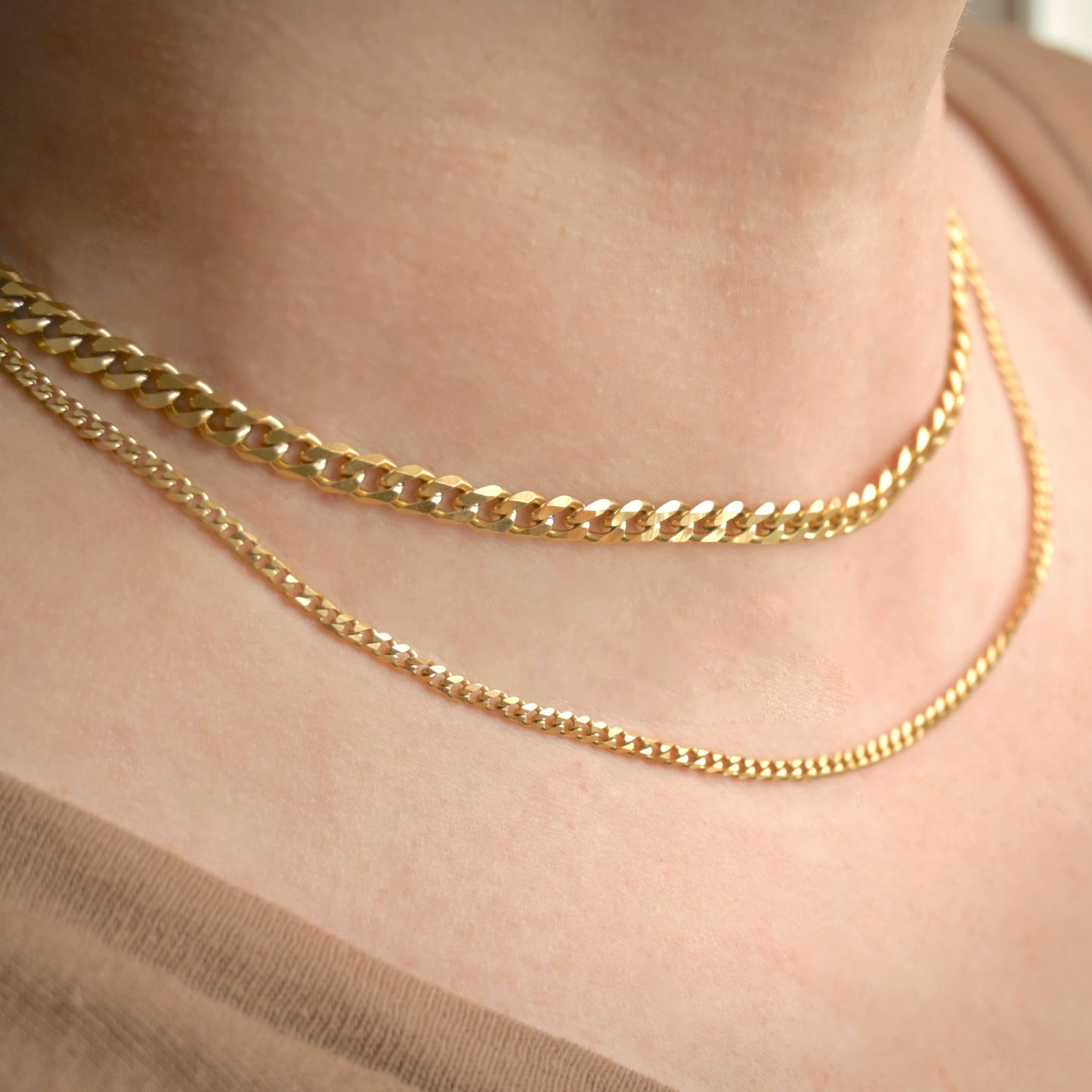 Tw gold curb chain necklaces, one 14-inch with 5mm links, one 16-inch with 3mm links, on a person's neck with a neutral background