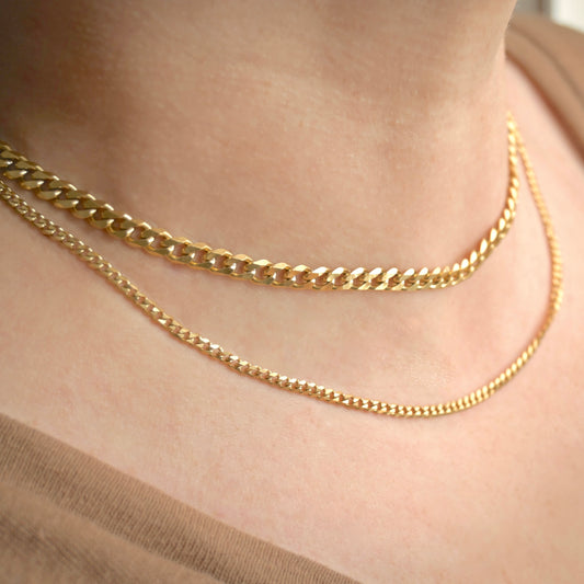 Tw gold curb chain necklaces, one 14-inch with 5mm links, one 16-inch with 3mm links, on a person's neck with a neutral background