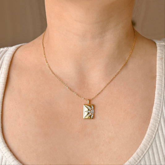 A close-up of a person's neck and chest wearing a delicate gold chain necklace with a small geometric pendant featuring a central clear stone.